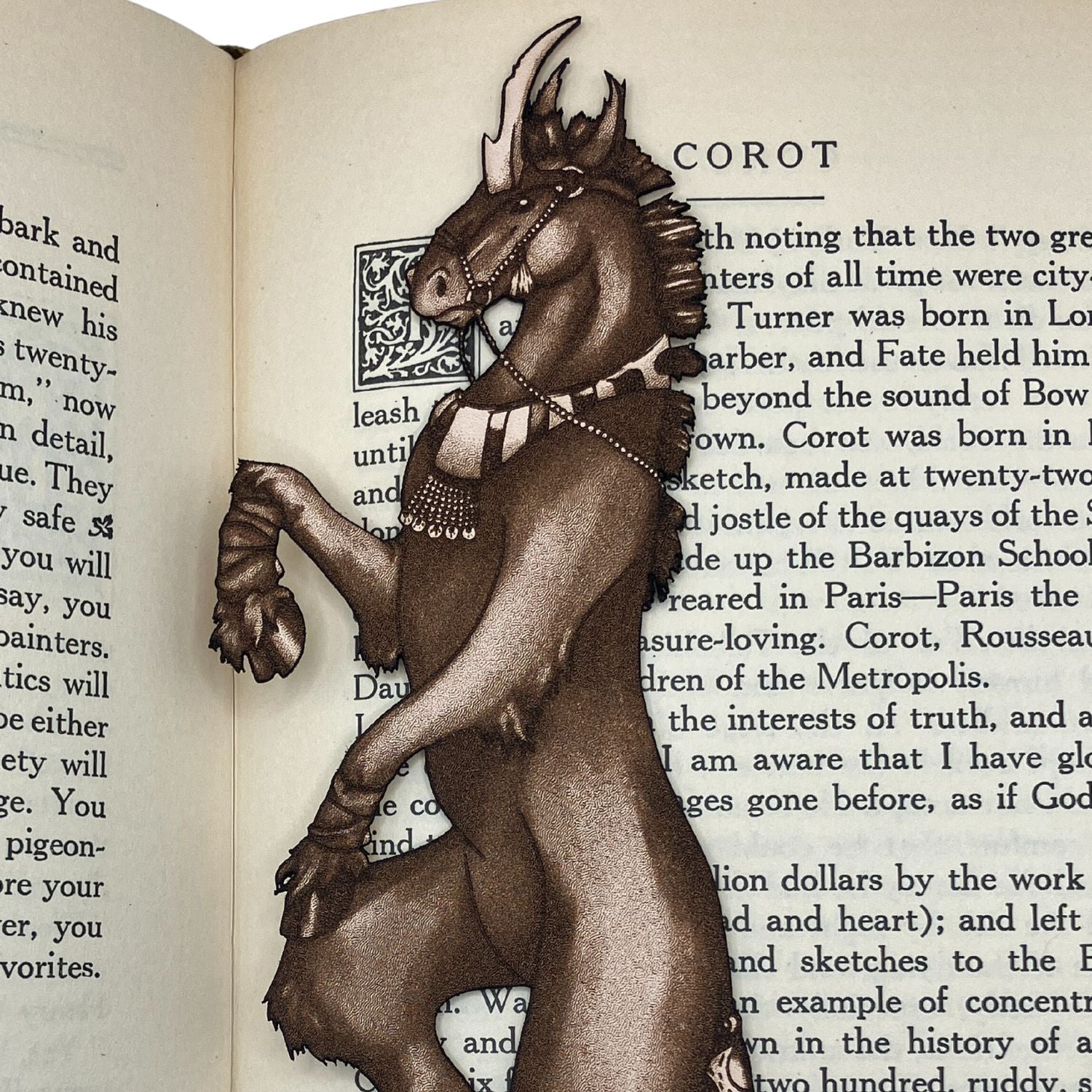 A leather bookmark shaped like a unicorn resting on the pages of an open book. The leather unicorn bookmark shows that the unicorn is wearing a collar, harness or bridle, trail cuff and leg wrappings. Dark Unicorn Bookmark - Leather Bookmarks Raven King Crafts