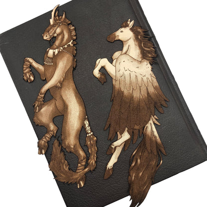 The unicorn bookmark is on the left and the Pegasus bookmark is on the right. Both bookmarks are resting on the cover of a closed leather book. Dark Unicorn Bookmark - Leather Bookmarks Raven King Crafts