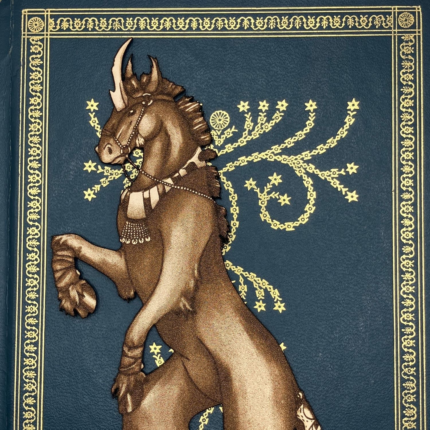 A close up of the head and chest of the unicorn bookmark. The bookmark is resting on the cover of a closed book. The book cover is blue with ornate gold decorations. Dark Unicorn Bookmark - Leather Bookmarks Raven King Crafts