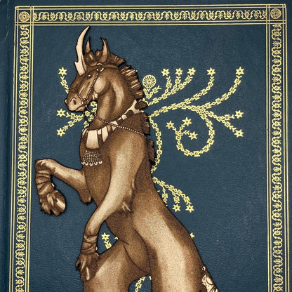 A close up of the head and chest of the unicorn bookmark. The bookmark is resting on the cover of a closed book. The book cover is blue with ornate gold decorations. Dark Unicorn Bookmark - Leather Bookmarks Raven King Crafts
