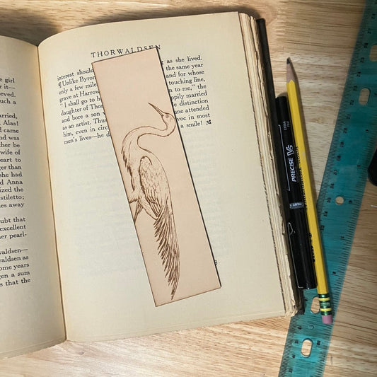 The photograph shows a leather bookmark with the art of an egret bird engraved on it. The leather bookmark is sitting on the pages of an open book.