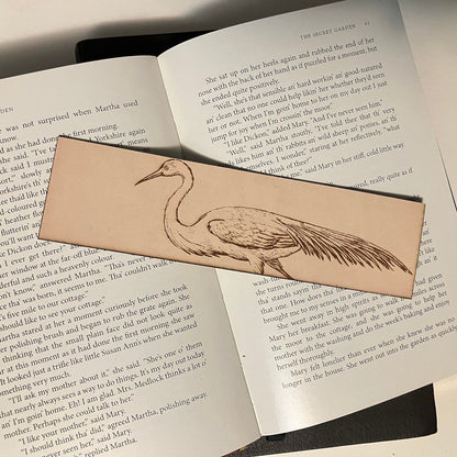 The photograph shows a leather bookmark with the art of an egret bird engraved on it. The leather bookmark is sitting on the pages of an open book. Egret Bookmark - Leather Bookmarks Raven King Crafts