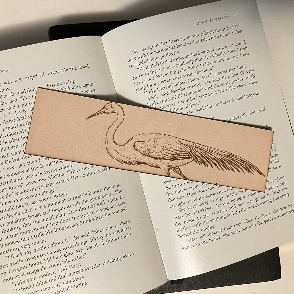 The photograph shows a leather bookmark with the art of an egret bird engraved on it. The leather bookmark is sitting on the pages of an open book.