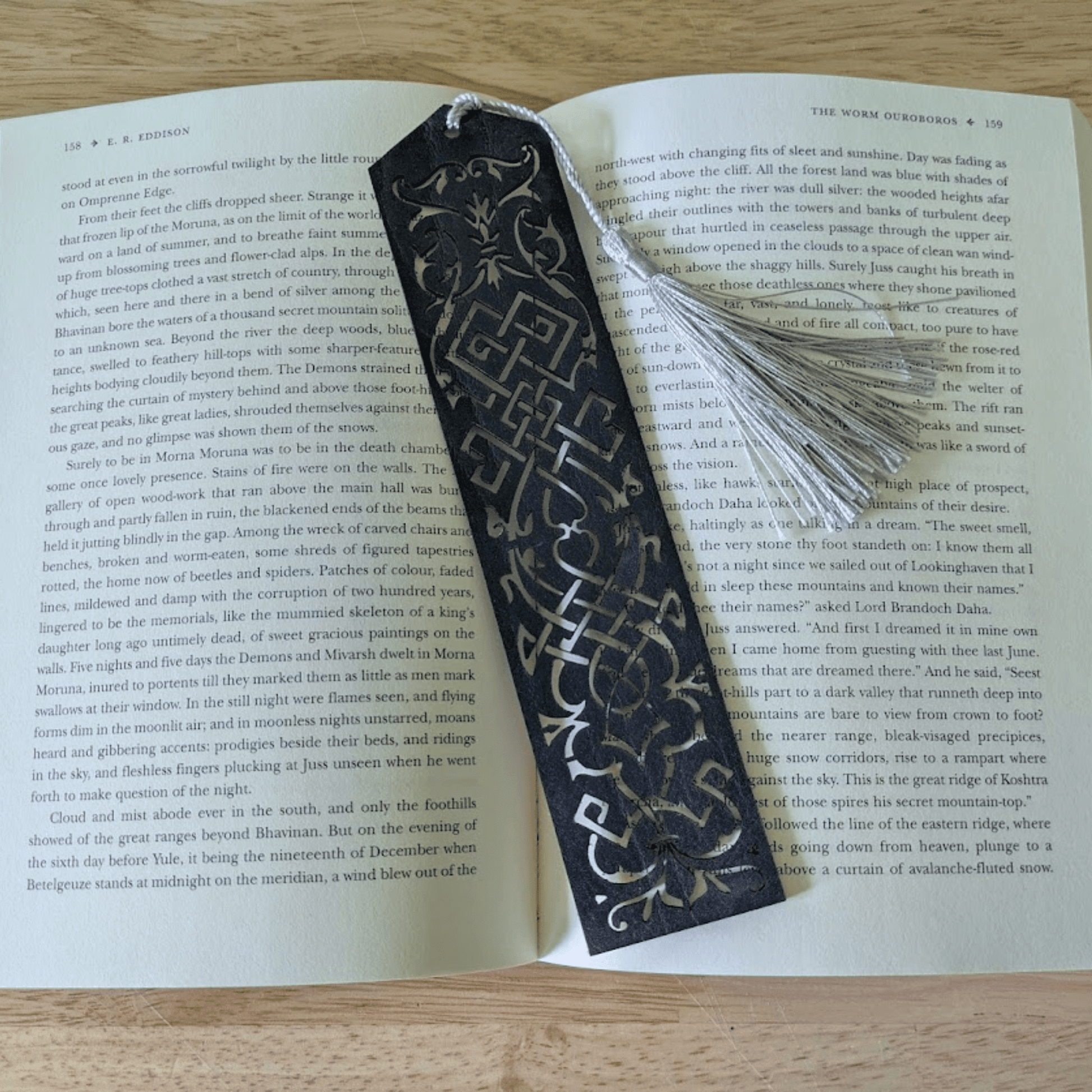 A dark blue leather bookmark resting on the pages of an open book. Blue Cutout Leather Bookmark with Tassel Hole Bookmarks Raven King Crafts Elvish Style Leather Bookmarks - Pick your color and your tassel Bookmarks Raven King Crafts