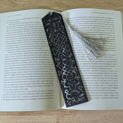 A dark blue leather bookmark resting on the pages of an open book. Blue Cutout Leather Bookmark with Tassel Hole Bookmarks Raven King Crafts Elvish Style Leather Bookmarks - Pick your color and your tassel Bookmarks Raven King Crafts