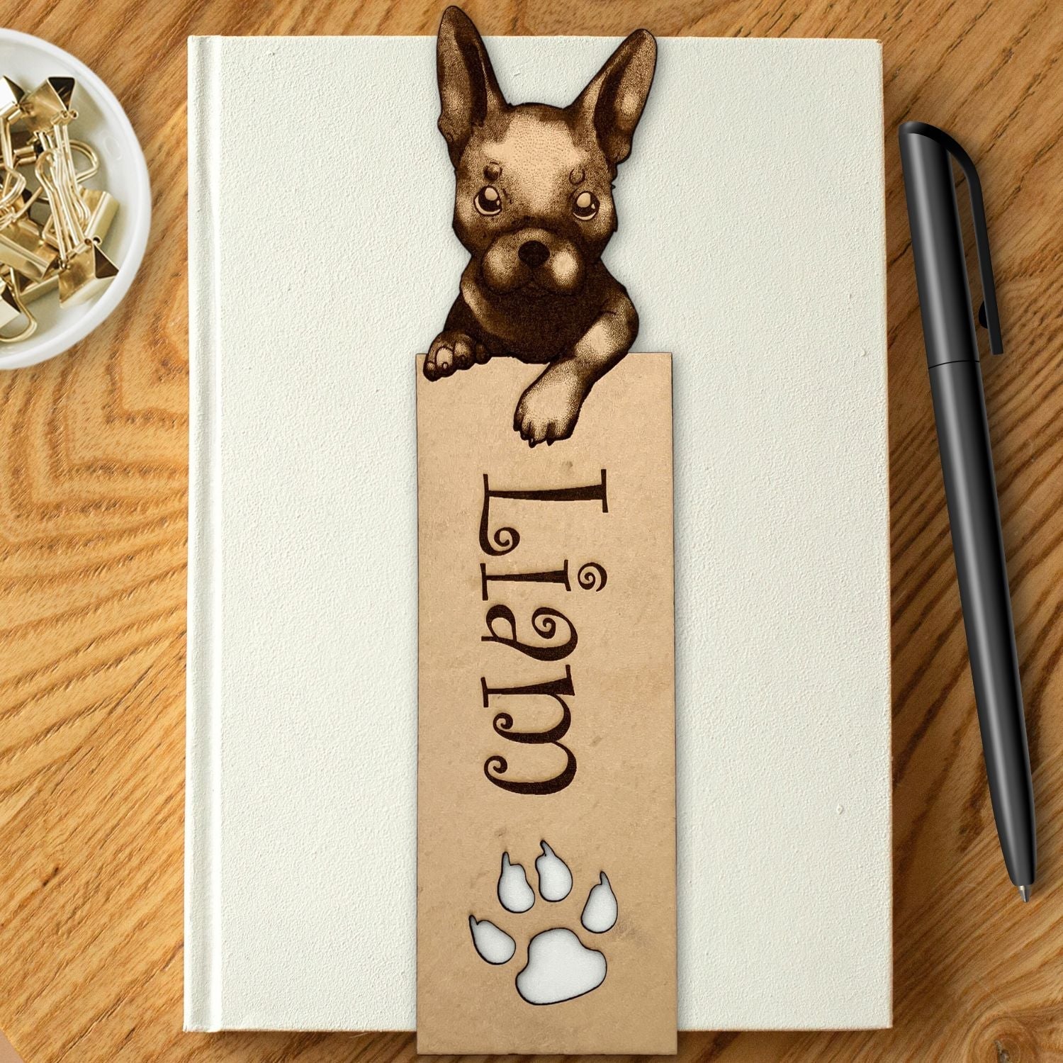 A french bulldog bookmark personalized with the name Liam. It is resting on the cover of a white book. The book is resting on a wood table, with a pen to the right and a bowl with clips in it to the left. French Bulldog Bookmark Bookmarks Raven King Crafts