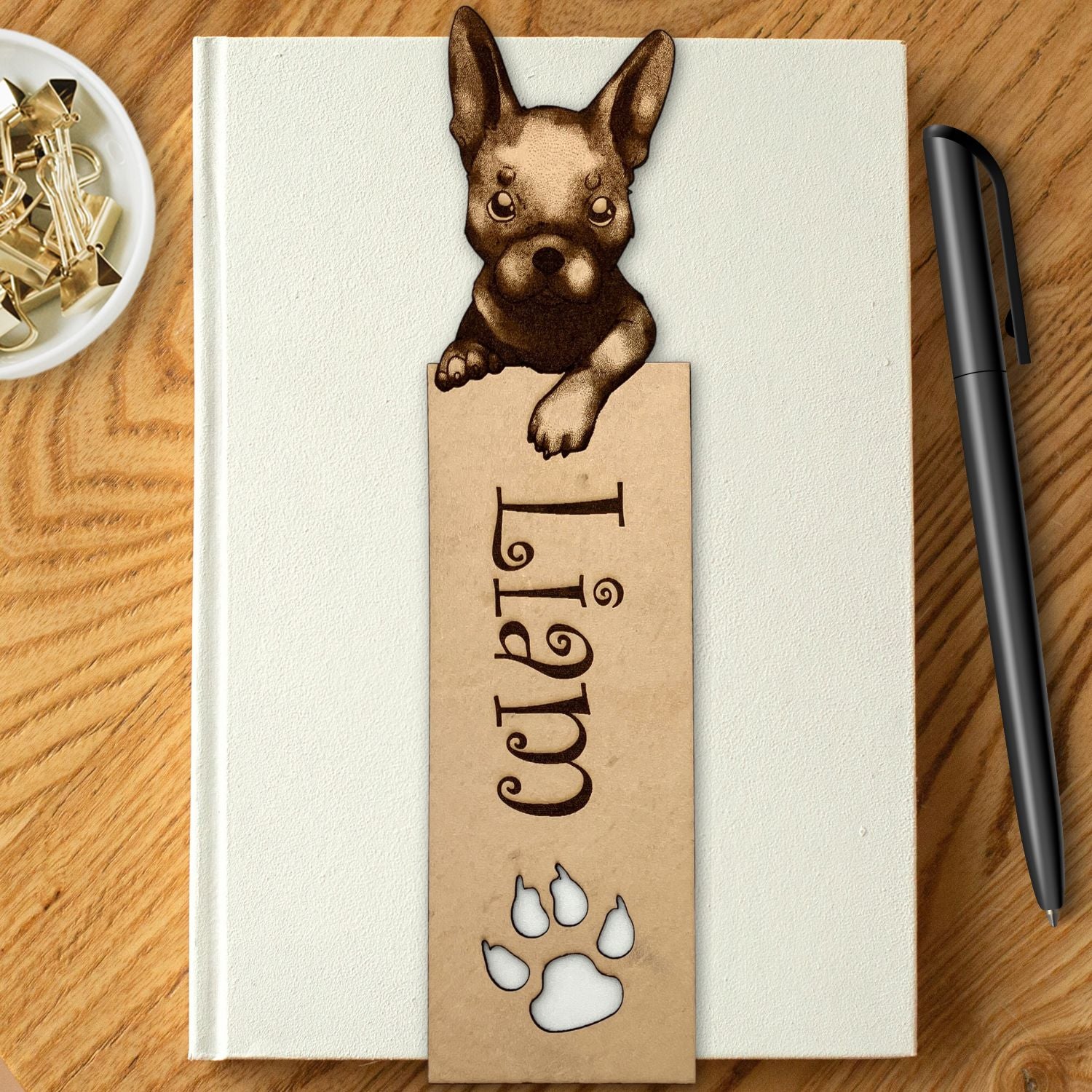 A french bulldog bookmark personalized with the name Liam. It is resting on the cover of a white book. The book is resting on a wood table, with a pen to the right and a bowl with clips in it to the left.