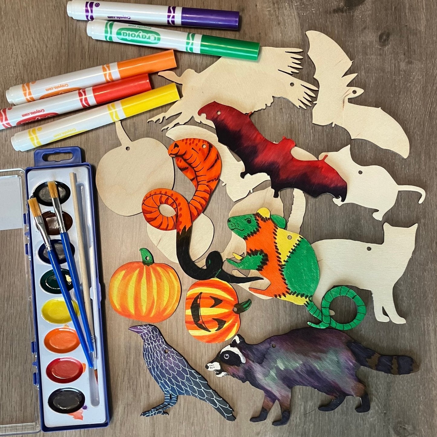 Photo of the Halloween Ornament Blank set with some of the ornament colored. It also shows the watercolor set and some markers that you can add to your set to make it a full kit. The decorated examples are an orange snake, red and black bat, zombie rat, pumpkins, raccoon and crow.