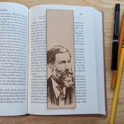 A leather rectangular bookmark resting on the pages of an open book. The art engraved on the bookmark is that of a handsome man with a well groomed beard and mustache. He is also wearing a suit and tie. The bookmark is resting on the pages of an open book.