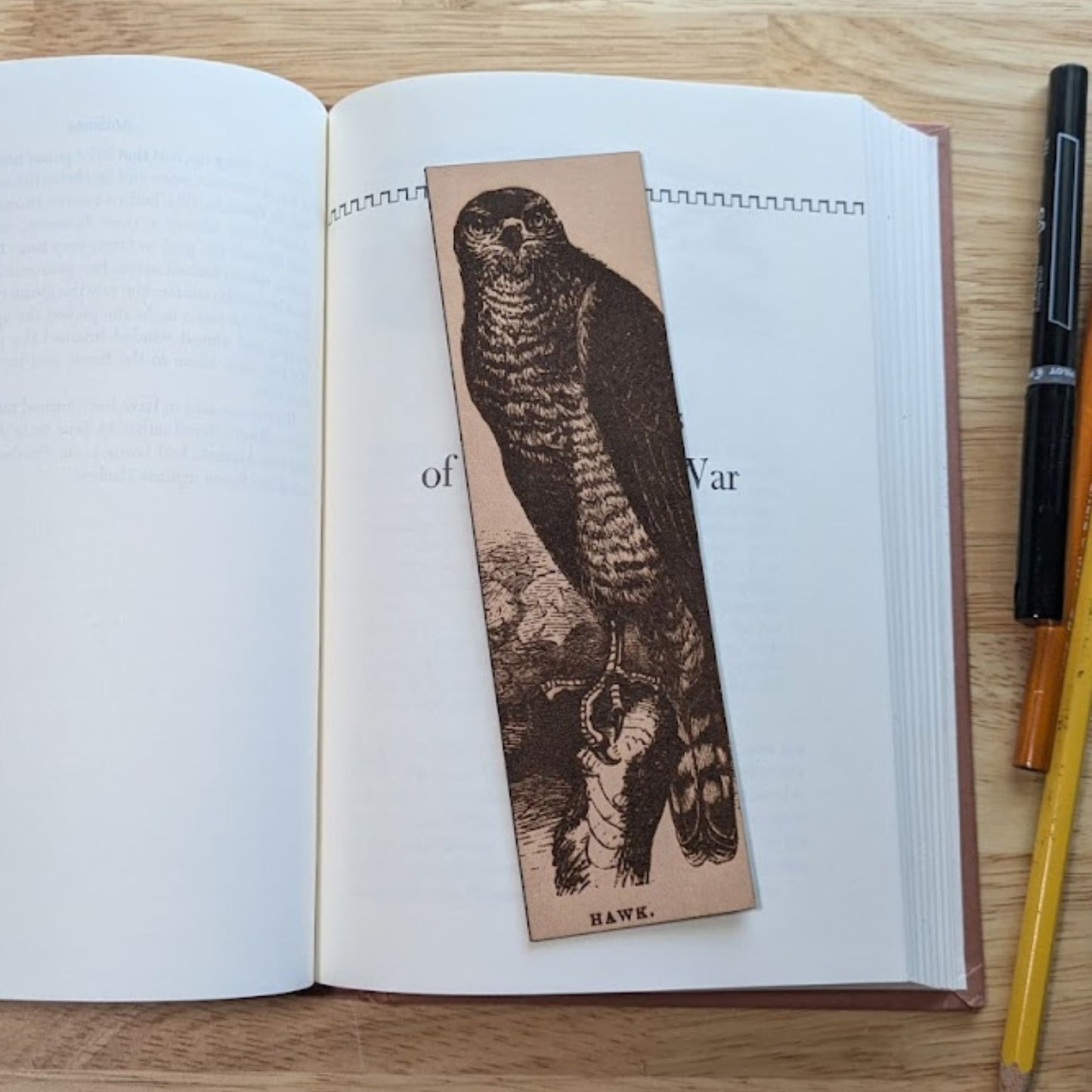 A rectangular leather bookmark resting on the pages of an open book. The antique art of a hawk is engraved on the bookmark. 