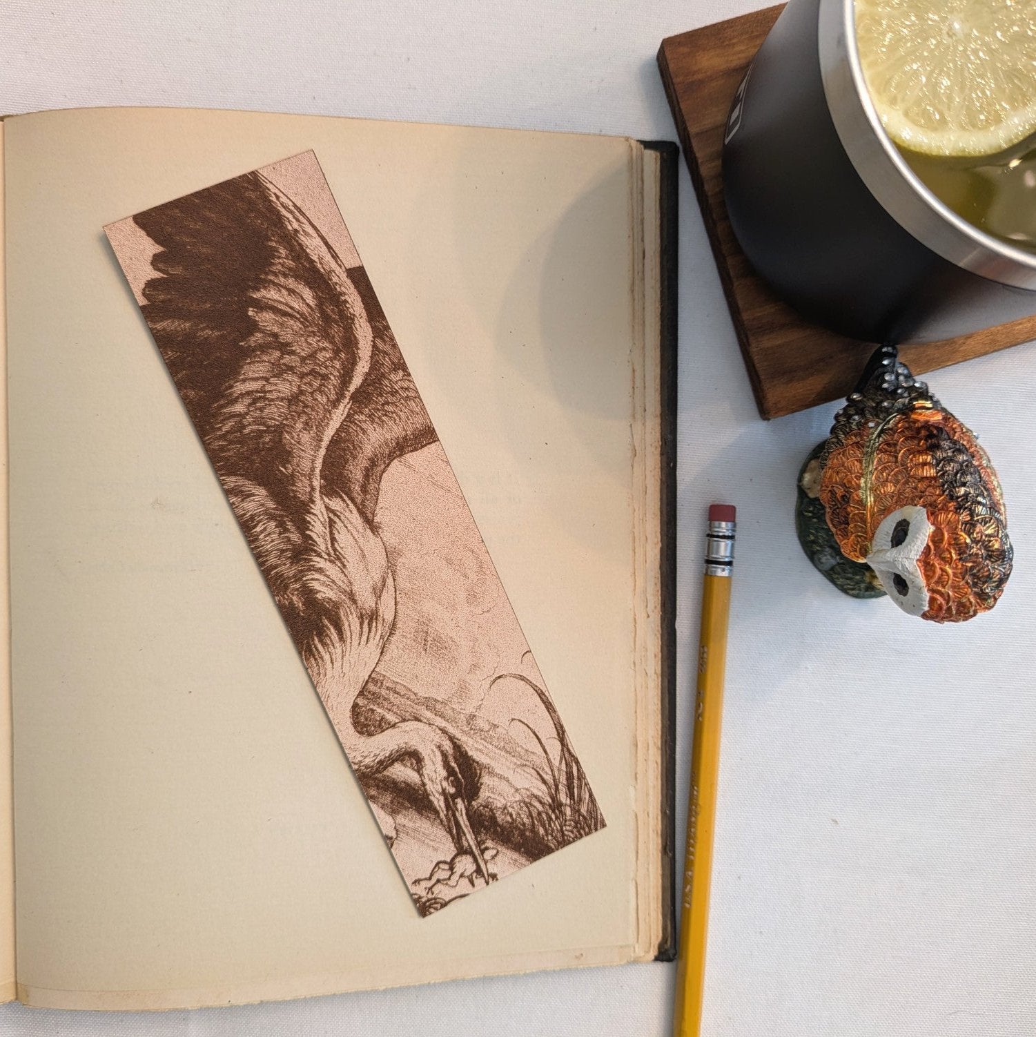 A photograph of the heron with a fog bookmark. The leather bookmark is resting on the page of an open book. Also in hte photo is a drink, owl accessory and pencil.