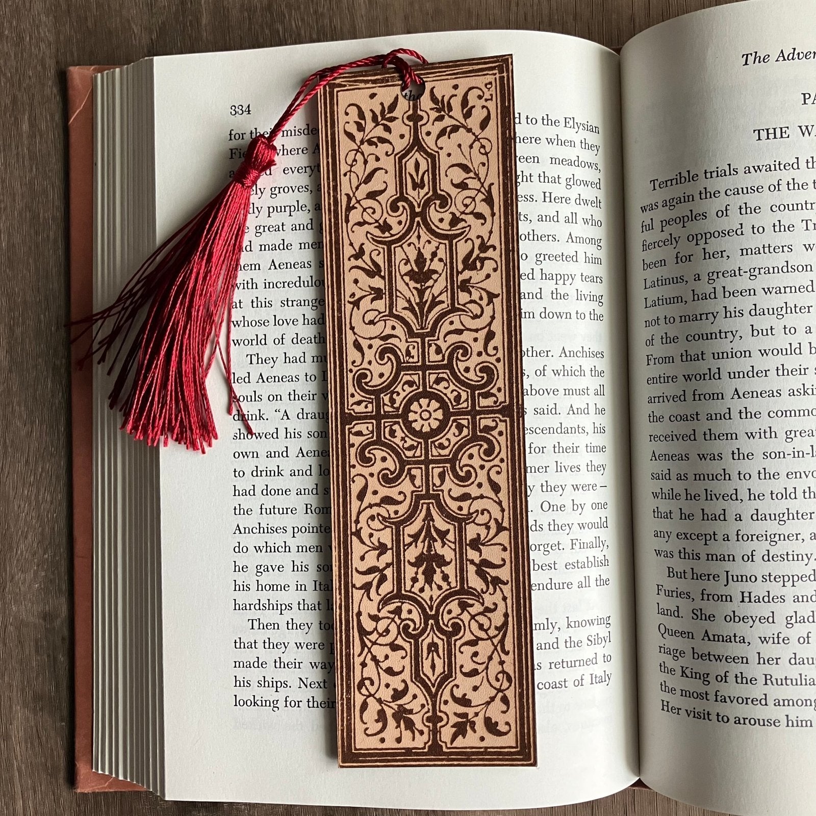 A photograph of a rectangular leather bookmark with an ornate design engraved on it. The bookmark in this pictures has a red tassel and is sitting on an open book.