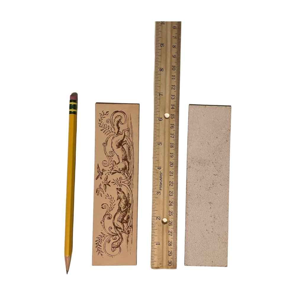The rectangular leather bookmark with the historical illustration of 2 foxes trying to catch birds engraved on it. Next to the leather fox bookmark are a pencil and a ruler to help show scale. To the right is the back of the bookmark, which is unfinished. Historical Fox Illustration Bookmark Bookmarks Raven King Crafts