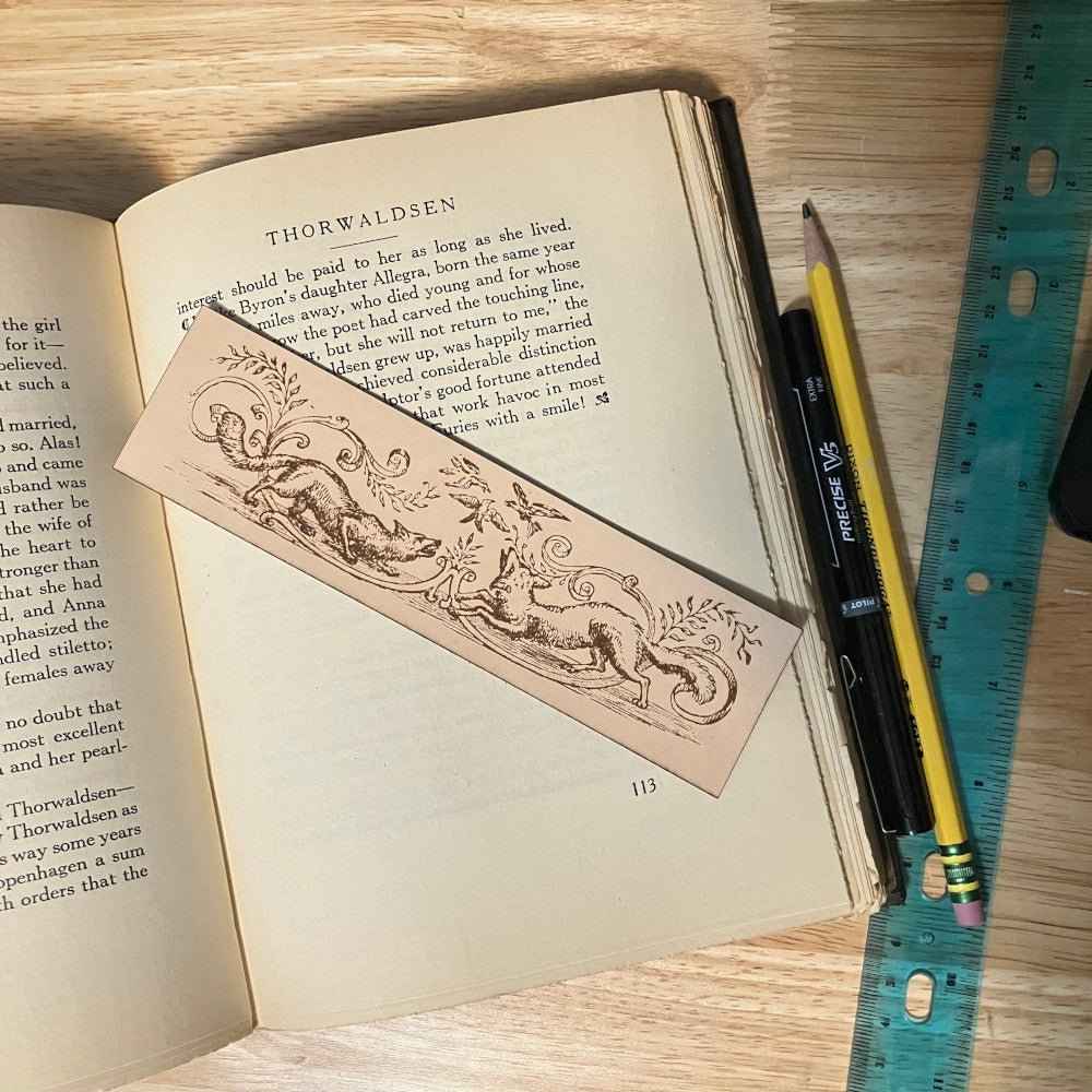 A rectangular leather bookmark with the historical art of 2 foxes trying to catch birds engraved on it. The leather fox bookmark is resting on the pages of an open book. Historical Fox Illustration Bookmark Bookmarks Raven King Crafts
