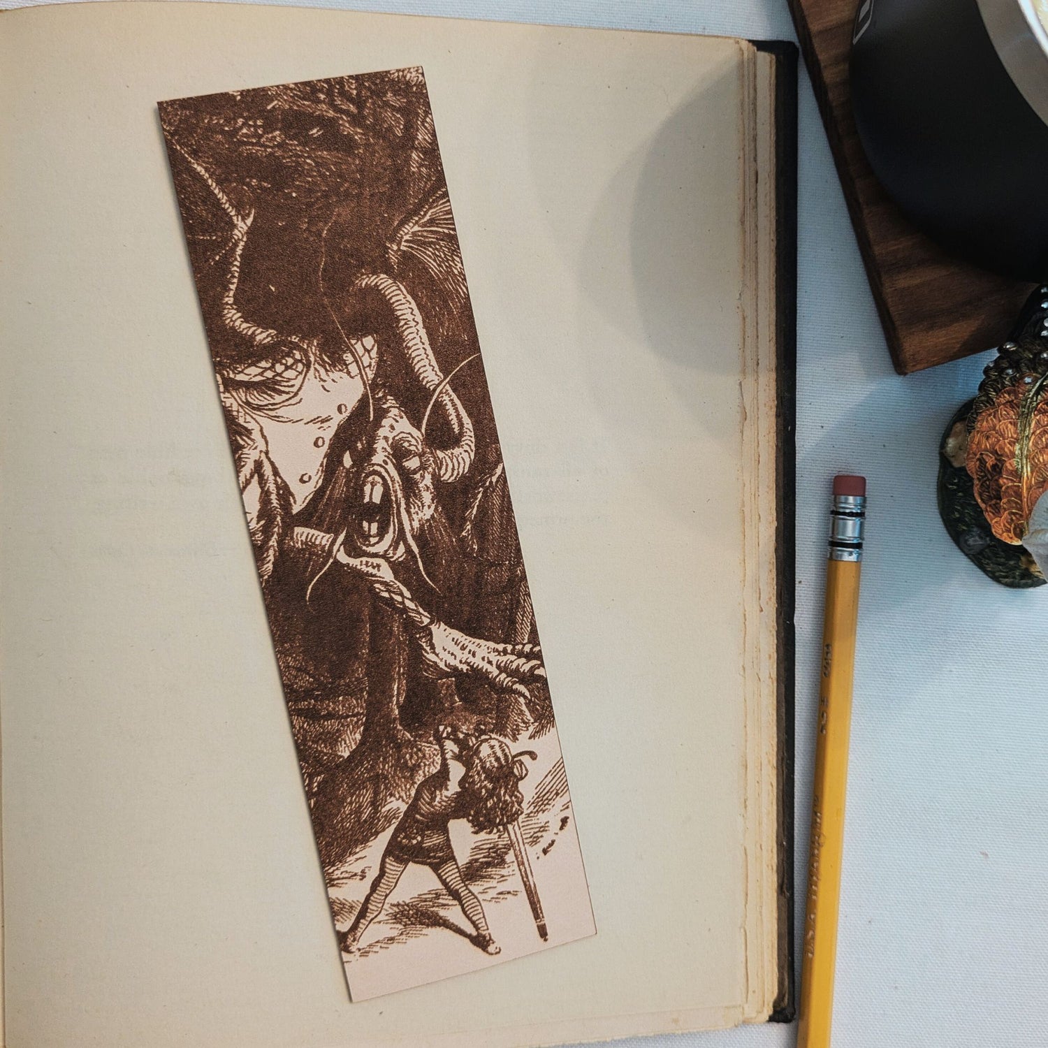 The photograph is of a rectangular leather bookmark with the antique art of a jabberwocky and young man in battle. The jabberwock art is engraved into the leather. The leather jabberwocky bookmark is resting on the pages of an open book. A pencil is to the right of it for scale. 