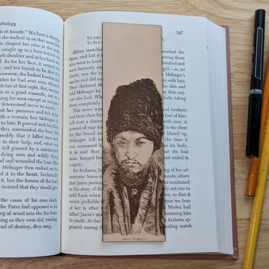 A leather rectangular bookmark with the historical art of the Khan of Khiva engraved on it. The Khan is a well dressed man with a beard. The bookmark is resting on the pages of an open book.