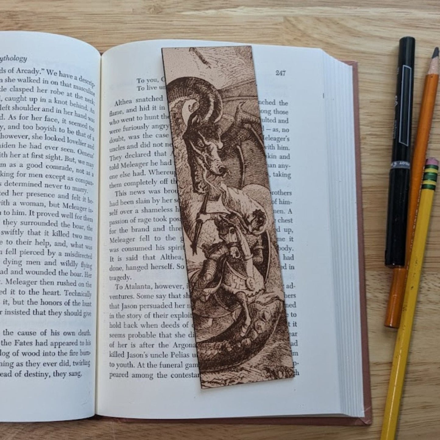 A photograph of a rectangular leather bookmark with the art of dragon fighting a knight engraved on it. The bookmark itself is resting on the pages of an open book. The knight in the art is dressed in armor and riding his horse. The dragon is much larger than the knight. Knight Fighting a Dragon - Leather Historical Bookmark Bookmarks Raven King Crafts Knight Fighting a Dragon - Leather Historical Bookmark Bookmarks Raven King Crafts