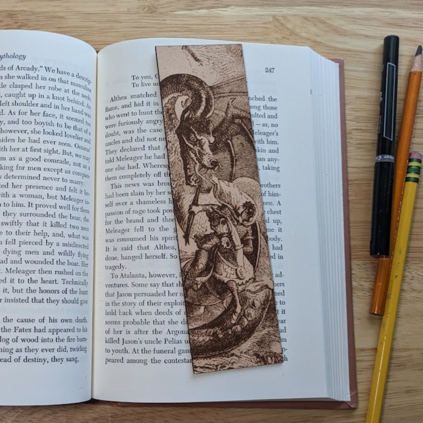 A photograph of a rectangular leather bookmark with the art of dragon fighting a knight engraved on it. The bookmark itself is resting on the pages of an open book. The knight in the art is dressed in armor and riding his horse. The dragon is much larger than the knight. 