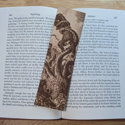A photograph of the rectangular leather bookmark with the art of a kraken attacking a salior engraved on it. The bookmark itself is resting on the pages of an open book. Kraken (Giant Octopus) Leather Bookmark Bookmarks Raven King Crafts Kraken (Giant Octopus) Leather Bookmark Bookmarks Raven King Crafts