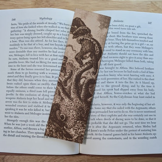 A photograph of the rectangular leather bookmark with the art of a kraken attacking a salior engraved on it. The bookmark itself is resting on the pages of an open book.