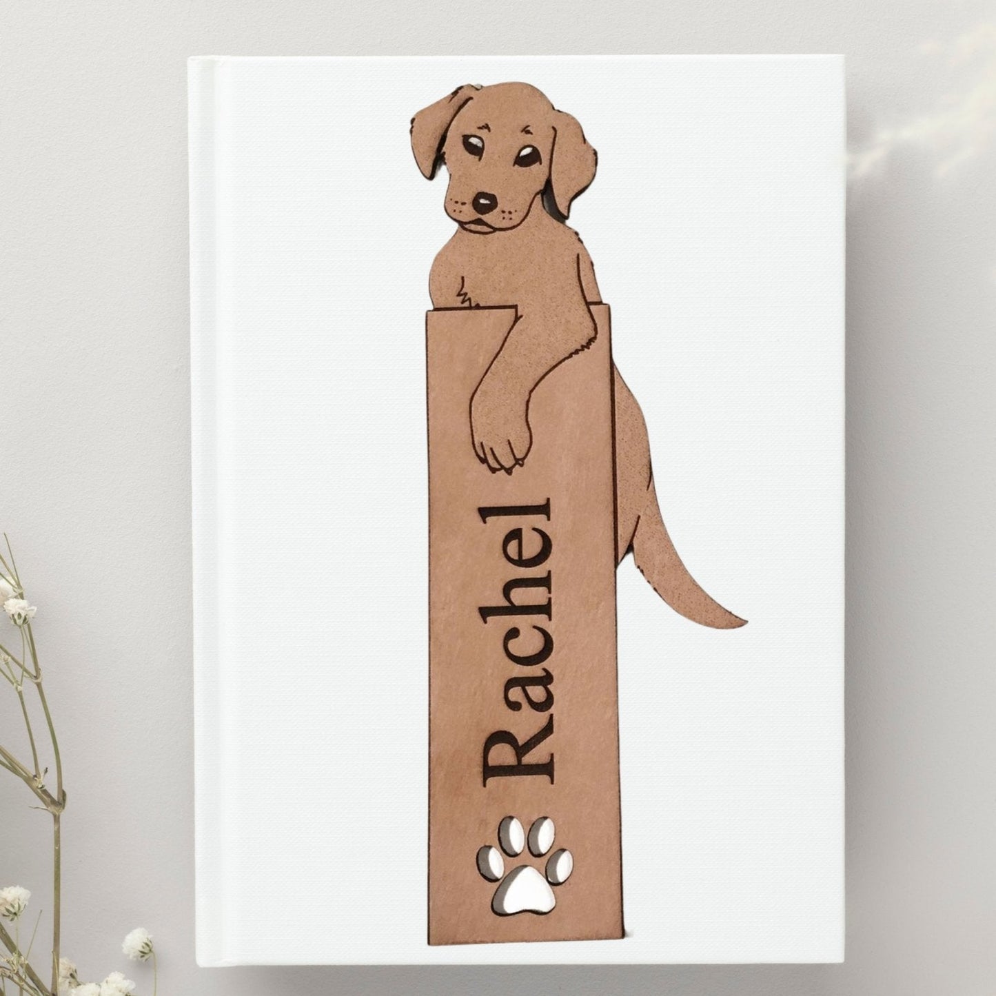 A bookmark of a Labrador retriever puppy. You can get this bookmark engraved to personalize it with a name or short message. The bookmark is resting on the cover of white book.