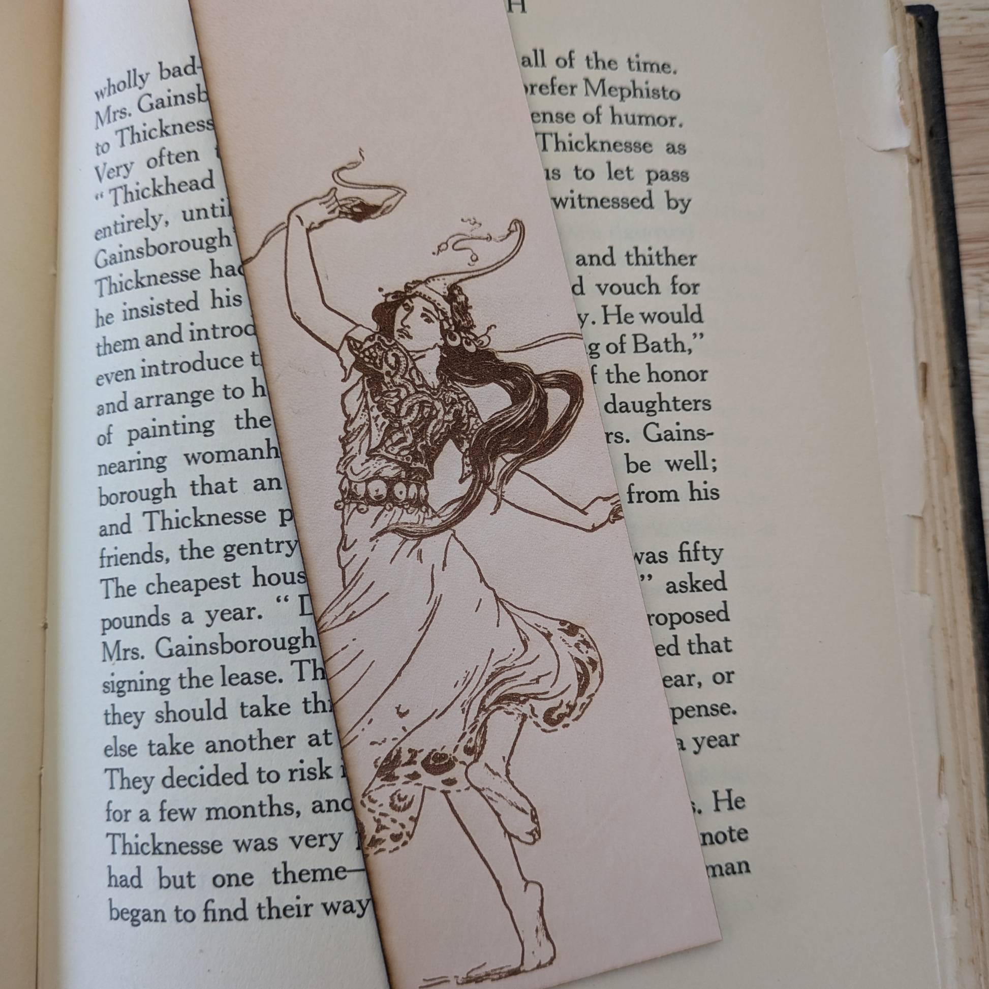 The leather bookmark with the historical art of a woman dancing engraved on it. The bookmark is resting on the pages of an open book. 