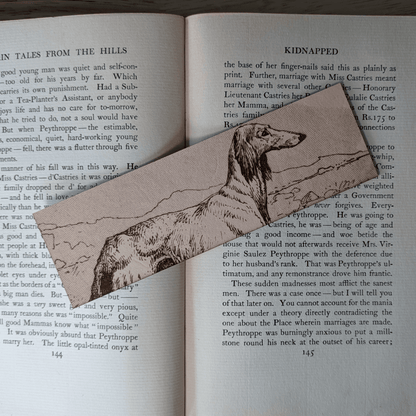 A rectangular leather bookmark with the historical art of an Afghan Greyhound dog engraved on it. The bookmark is resting on the pages of an open book. Leather Afghan Greyhound Bookmark Bookmarks Raven King Crafts Leather Afghan Greyhound Bookmark Bookmarks Raven King Crafts