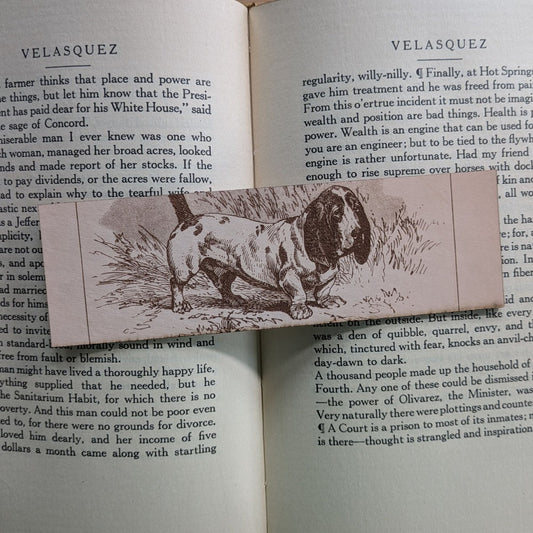 A rectangular leather bookmark with the historical art of a basset hound dog engraved on it. The bookmark is resting on the pages of an open book. Leather Basset Hound Bookmark Bookmarks Raven King Crafts Leather Basset Hound Bookmark Bookmarks Raven King Crafts