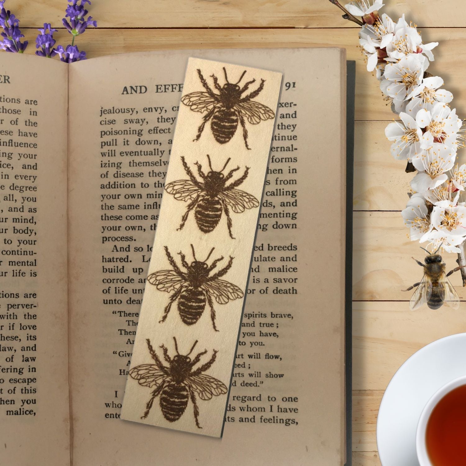 The leather bee bookmark resting on the pages of an open book. The leather bookmark has 4 bees engraved on it. To the right is a sprig of a plant with flowers and a bee. At the bottom right hand of the image is a tea cup.