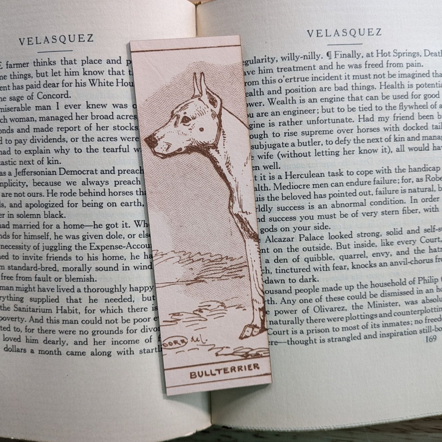 A rectangular leather bookmark with the historical art of a bull terrier dog engraved on it. The bookmark is resting on the pages of an open book. Leather Bull Terrier Dog Bookmark Bookmarks Raven King Crafts Leather Bull Terrier Dog Bookmark Bookmarks Raven King Crafts
