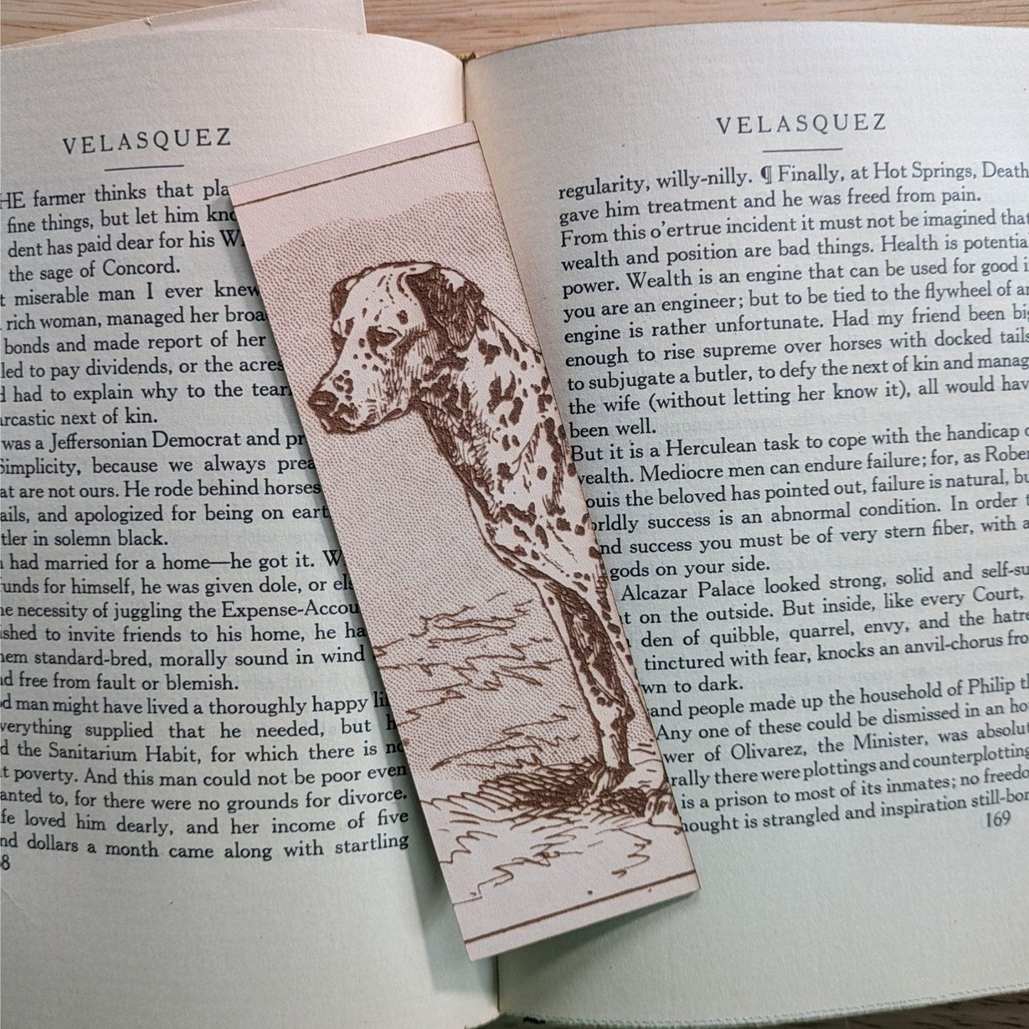 A rectangular leather bookmark with the historical art of a Dalmatian dog engraved on it. The bookmark is resting on the pages of an open book. Leather Dalmation Dog Bookmarks Bookmarks Raven King Crafts Leather Dalmatian Dog Bookmarks Bookmarks Raven King Crafts