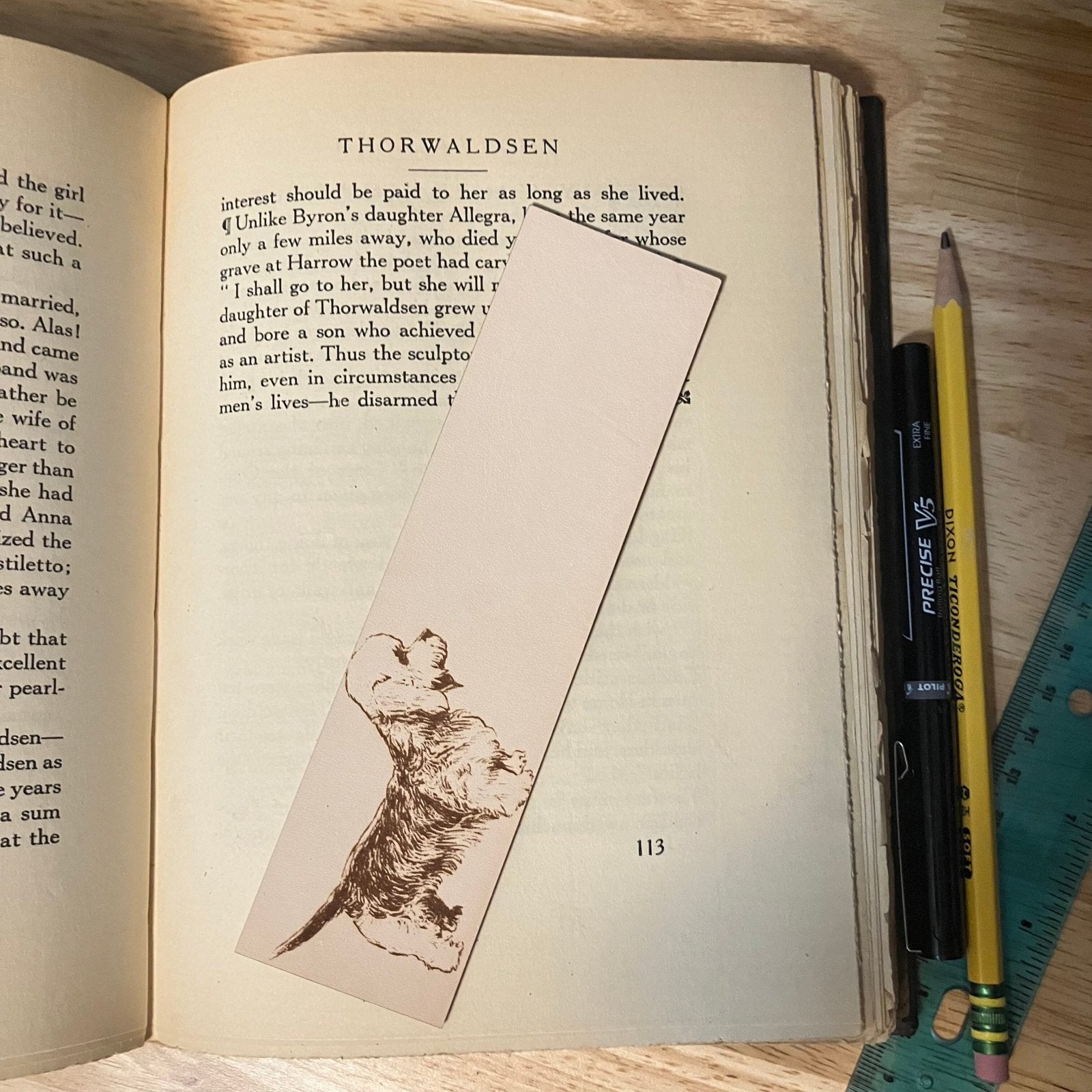 The leather Dandie Dinmont Terrier dog bookmark resting on the pages of an open book. To the right of it is a pencil and pen for scale. Leather Dandie Dinmont Terrier Bookmark (toy dog) Bookmarks Raven King Crafts Leather Dandie Dinmont Terrier Bookmark (toy dog) Bookmarks Raven King Crafts