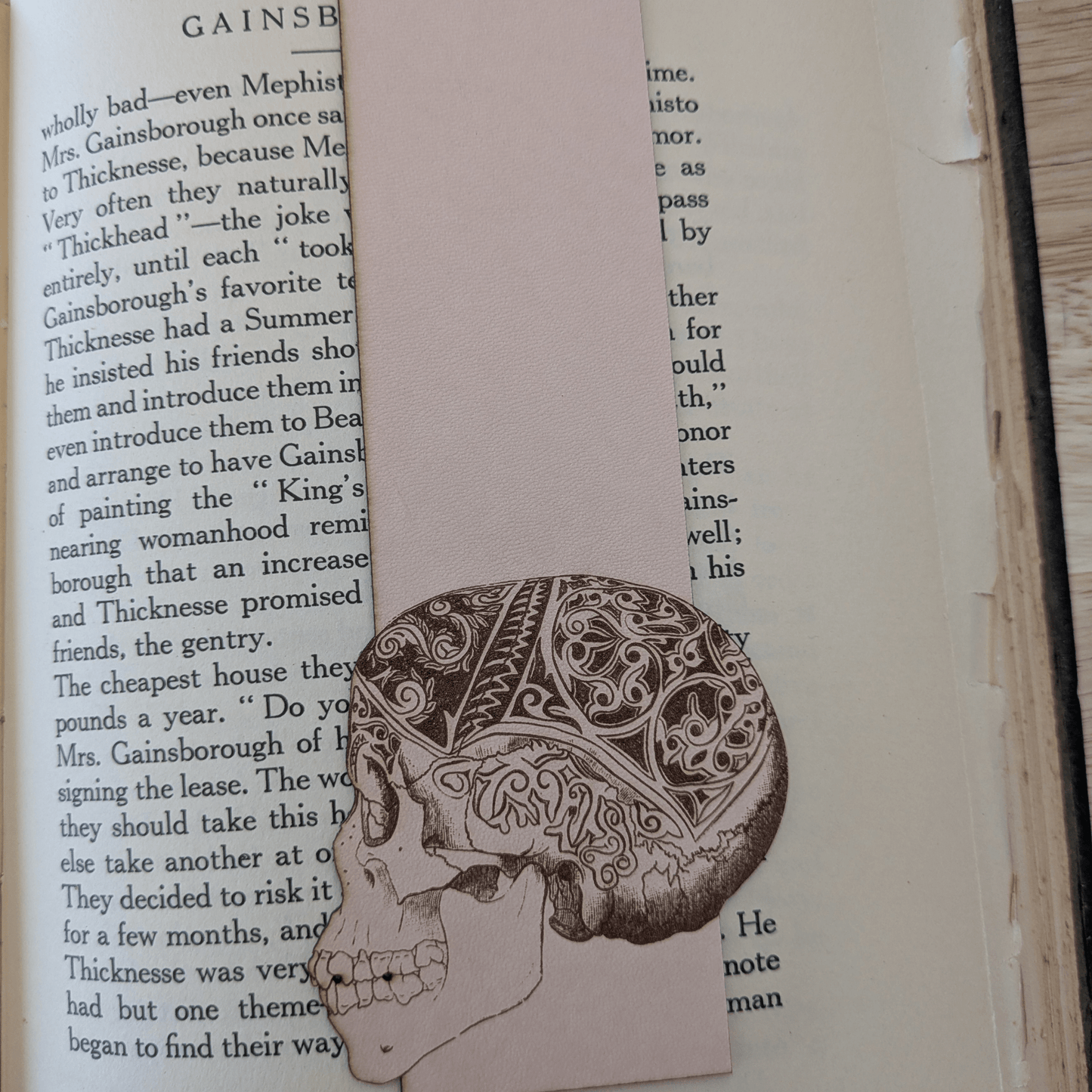 A leather bookmark engraved with the art of a skull on it. The skull bookmark is resting on the pages of an open book. Leather Engraved Skull Bookmark Bookmarks Raven King Crafts