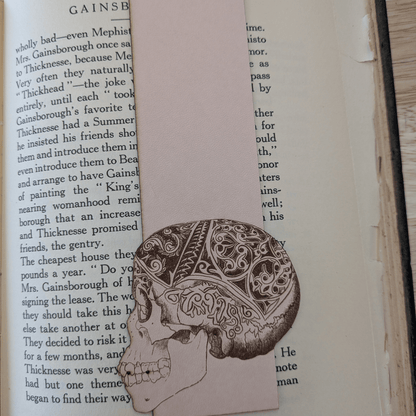 A leather bookmark engraved with the art of a skull on it. The skull bookmark is resting on the pages of an open book. Leather Engraved Skull Bookmark Bookmarks Raven King Crafts