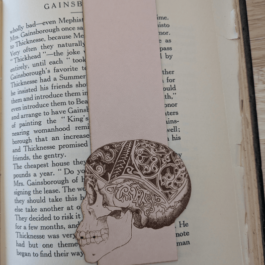 A leather bookmark engraved with the art of a skull on it. The skull bookmark is resting on the pages of an open book. Leather Engraved Skull Bookmark Bookmarks Raven King Crafts