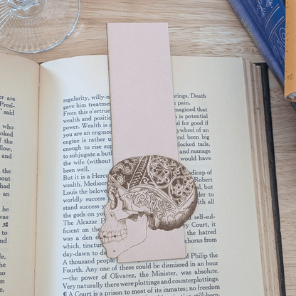 A leather bookmark engraved with the art of a skull on it. The skull bookmark is resting on the pages of an open book. Leather Engraved Skull Bookmark Bookmarks Raven King Crafts
