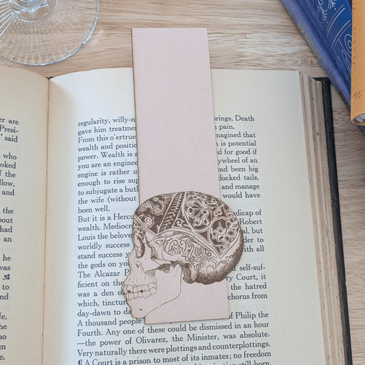 A leather bookmark engraved with the art of a skull on it. The skull bookmark is resting on the pages of an open book. Leather Engraved Skull Bookmark Bookmarks Raven King Crafts