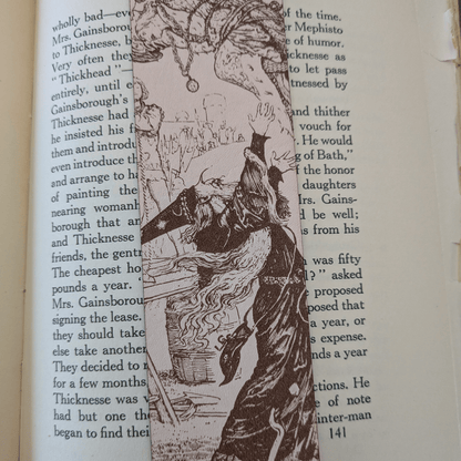 A leather bookmark engraved with the art of a wizard on it. The wizard bookmark is resting on the pages of an open book. Leather Engraved Wizard Bookmark Bookmarks Raven King Crafts