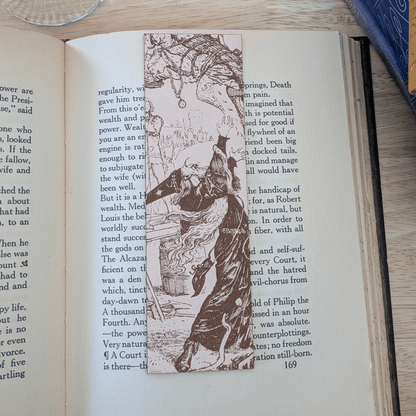 A leather bookmark engraved with the art of a wizard on it. The wizard bookmark is resting on the pages of an open book. Leather Engraved Wizard Bookmark Bookmarks Raven King Crafts