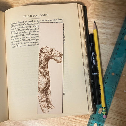 A rectangular leather bookmark with the historical art of an Irish Terrier engraved on it. The bookmark is resting on the pages of an open book. Leather Irish Terrier Bookmark Bookmarks Raven King Crafts Leather Irish Terrier Bookmark Bookmarks Raven King Crafts