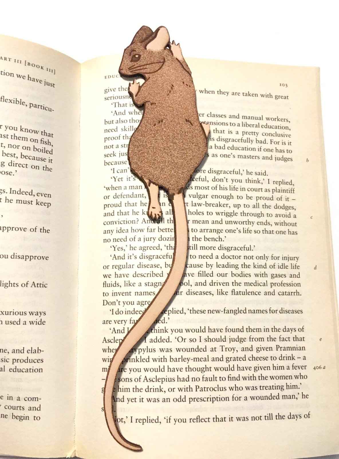 A photograph of exclusive climbing mouse leather bookmark resting on the pages of an open book. Leather Mouse Bookmark - Cute Little Book Buddy Bookmarks Raven King Crafts