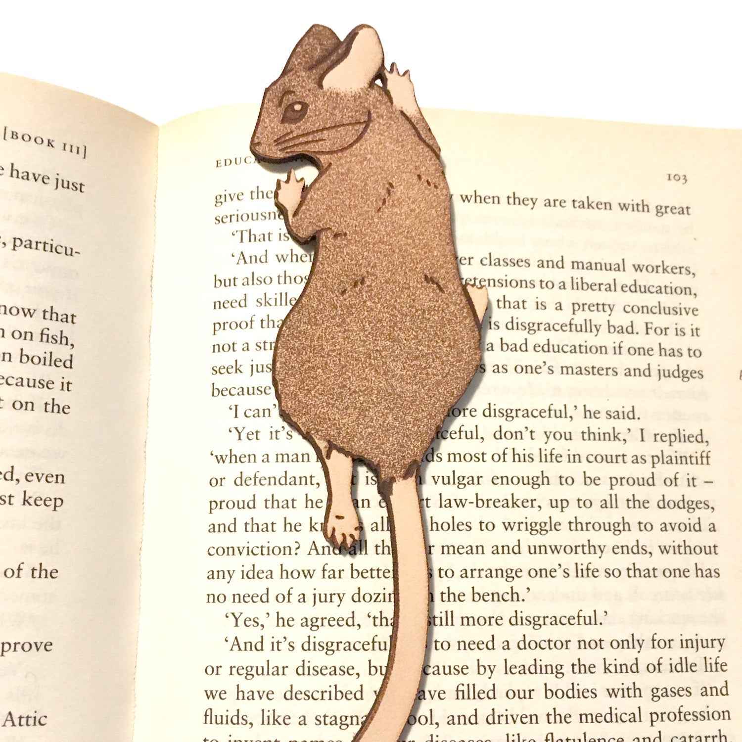 A photograph of exclusive climbing mouse leather bookmark resting on the pages of an open book. Leather Mouse Bookmark - Cute Little Book Buddy Bookmarks Raven King Crafts