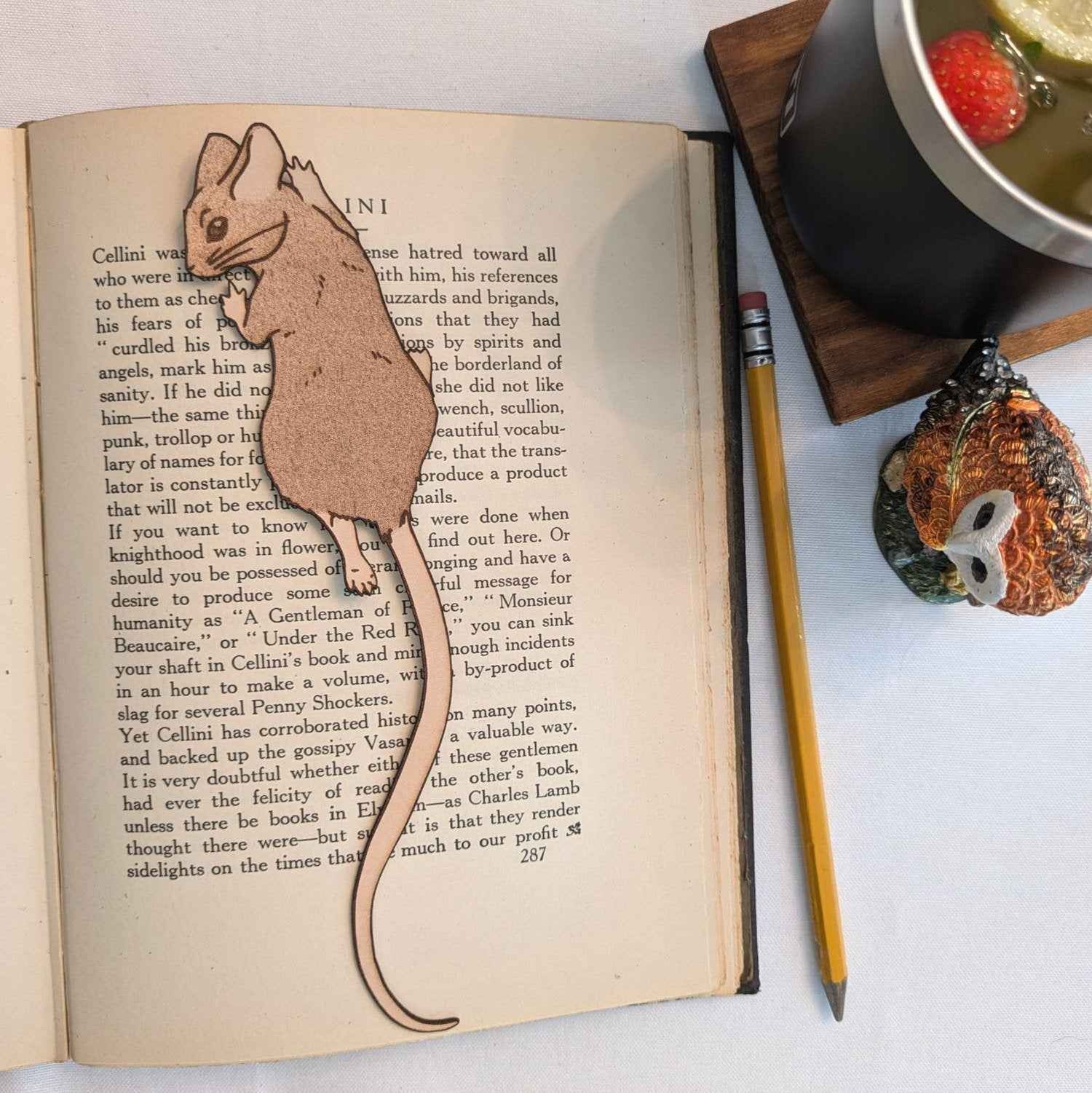 A photograph of exclusive climbing mouse leather bookmark resting on the pages of an open book. Leather Mouse Bookmark - Cute Little Book Buddy Bookmarks Raven King Crafts