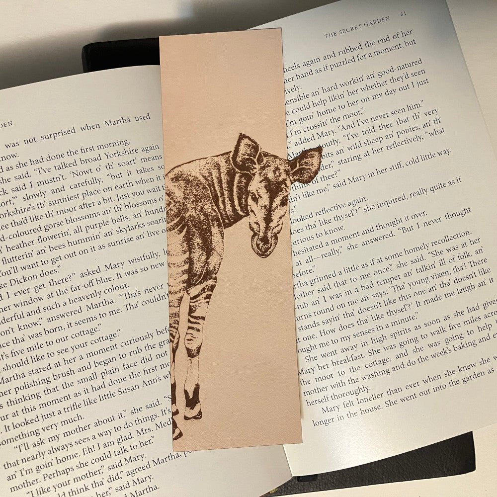 A leather bookmark with the art of an okapi engraved into it. The bookmark is resting on the pages of an open book.