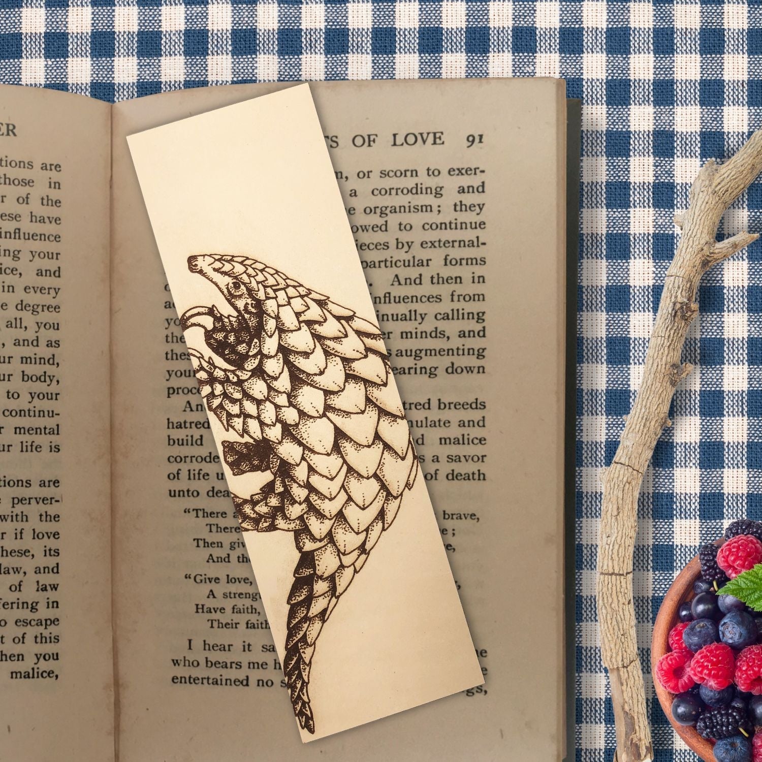 The leather pangolin bookmark resting on the open pages of a book. The book is resting on a blue checkered tablecloth. To the right of the book is a stick and a small bowl of berries.