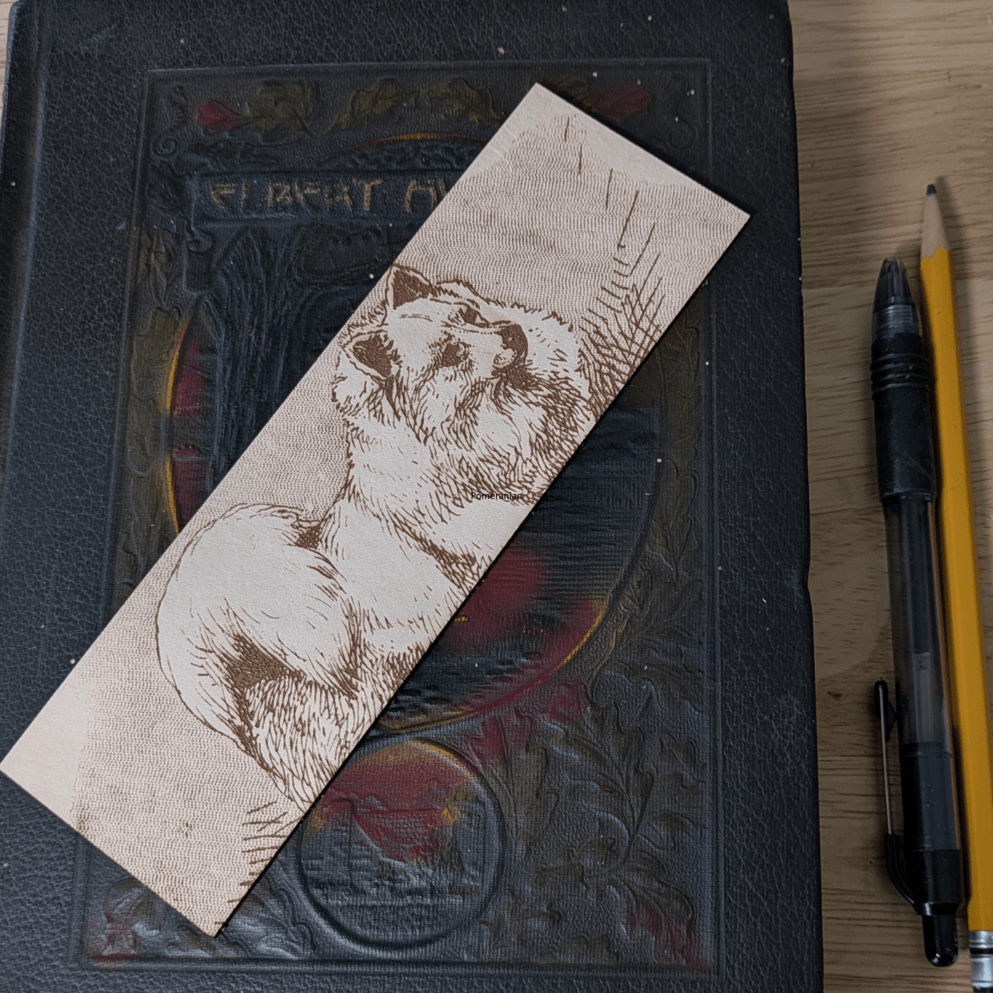 A rectangular leather bookmark with the historical art of a Pomeranian dog engraved on it. The bookmark is resting on the pages of an open book. Leather Pomeranian Bookmark Bookmarks Raven King Crafts Leather Pomeranian Bookmark Bookmarks Raven King Crafts