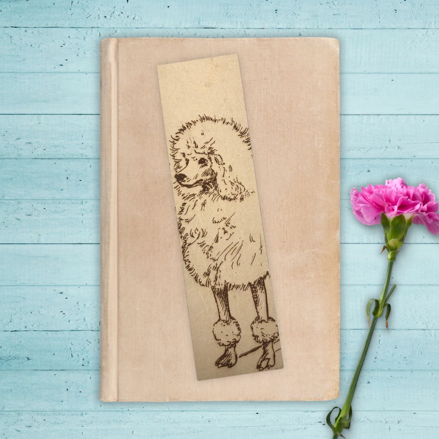 A leather bookmark of a standard poodle dog. The bookmark is sitting on the cover of a small closed tan book. The book is a wooden surface that appears to be painted a light blue.