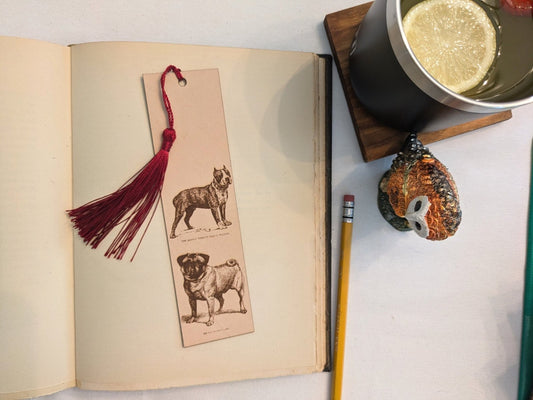 The leather Boston Terrier and Pug, dog bookmark resting on the pages of an open book. to the right of it is a pencil and a glass for scale. Leather Pug and Boston Terrier Bookmark (toy dog) Bookmarks Raven King Crafts Leather Pug and Boston Terrier Bookmark (toy dog) Bookmarks Raven King Crafts