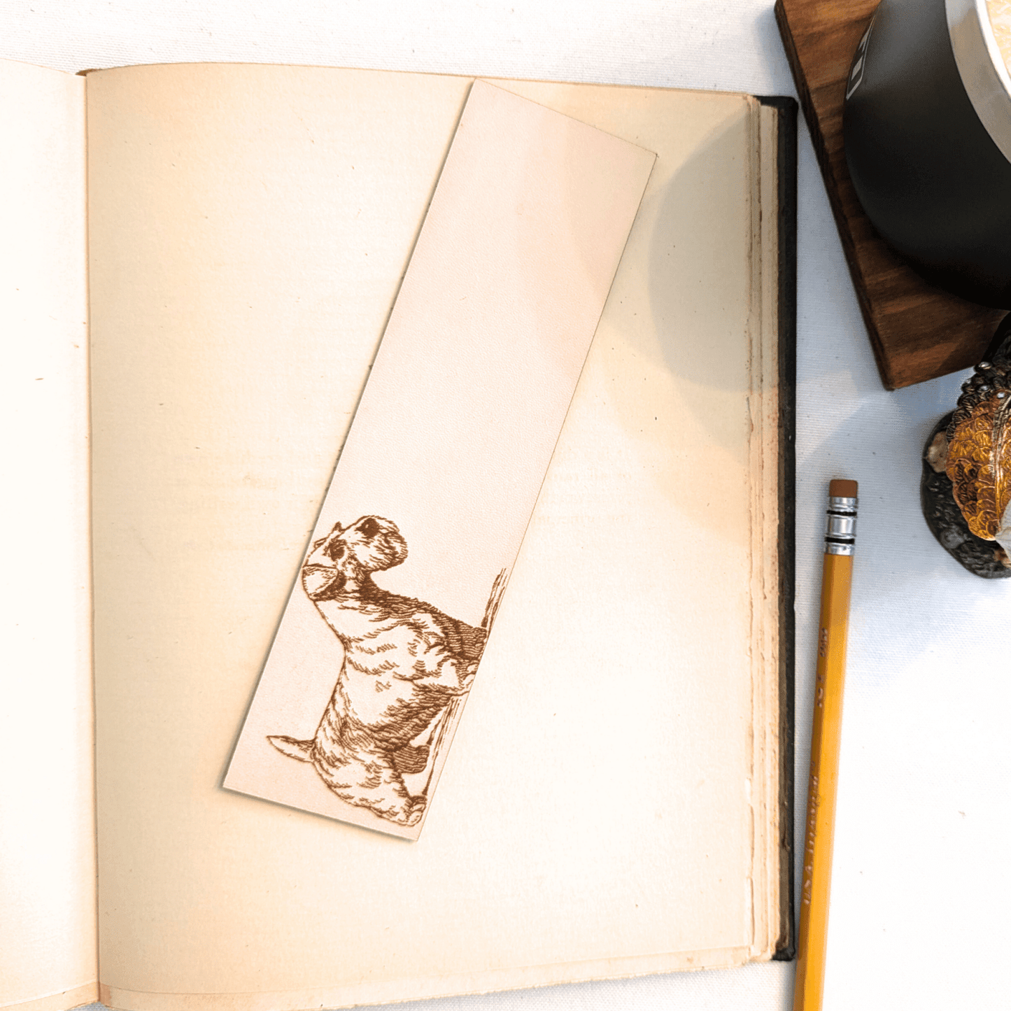 The leather Sealyham, dog bookmark resting on the pages of an open book. to the right of it is a pencil and a glass for scale. Leather Sealyham Terrier Bookmark (toy dog) Bookmarks Raven King Crafts Leather Sealyham Terrier Bookmark (toy dog) Bookmarks Raven King Crafts