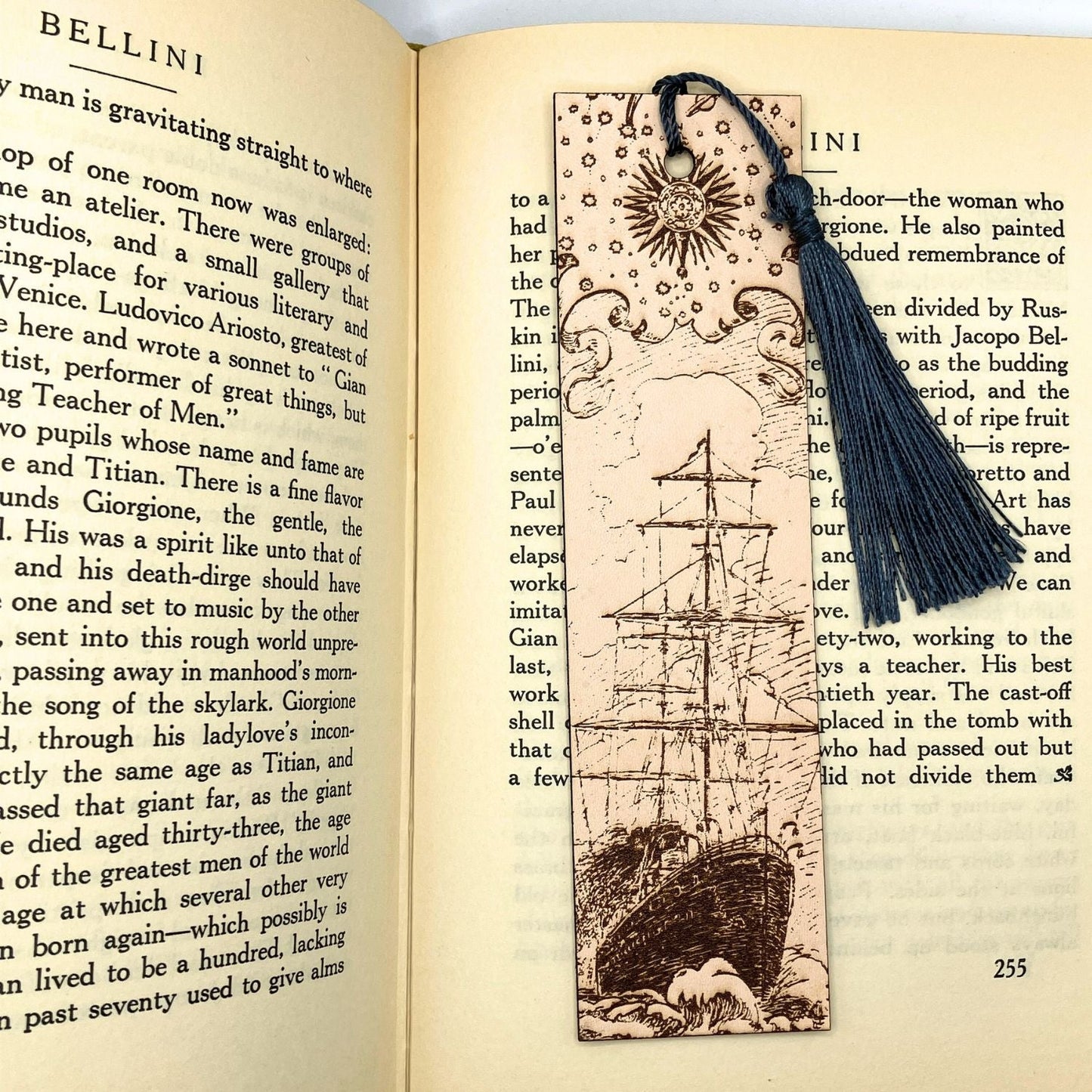 A leather bookmark with the historical art of a ship engraved onto it. This bookmark has a dark blue tassel and is resting on the pages of an open book. Leather Ship Bookmark Bookmarks Raven King Crafts